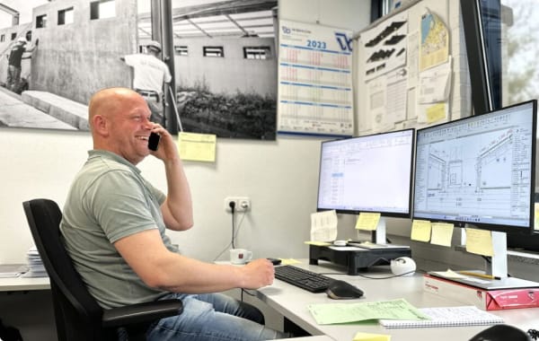 Medewerker werkt aan een bureau met twee schermen waarop bouwtekeningen en een planning zichtbaar zijn.