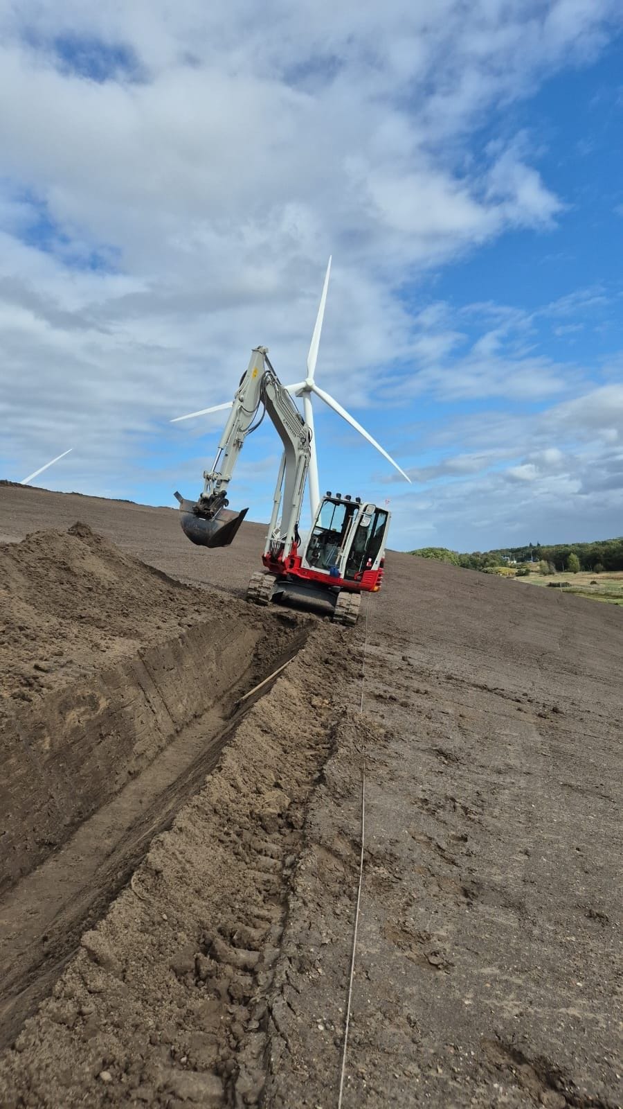 minigraver draait sleuf in zandige helling met windturbine op de achtergrond
